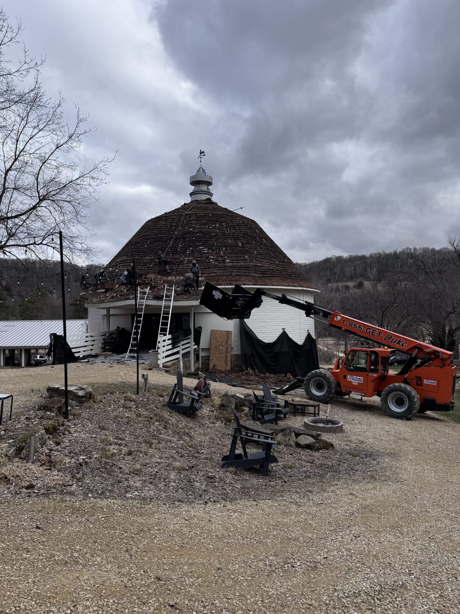 The round barn's original weathered roof mid-tear-off, dramatic stormy sky, Wacouta MN